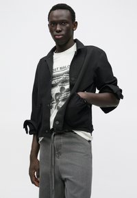 Čierna krátka bunda s gombíkmi, nosená cez bielu tričko s grafikou. Sivé džínsy s detailom na sťahovanie dopĺňajú outfit.