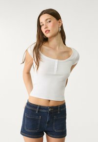 Vit, figurnära skjorta med rundad halsringning och femknappsstängning, parat med mörkblå denimshorts med synlig sömnad och fickor.