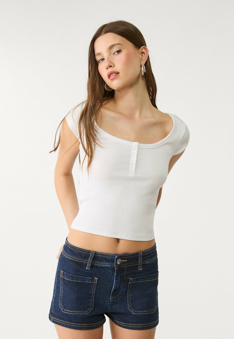 Vit, figurnära skjorta med rundad halsringning och femknappsstängning, parat med mörkblå denimshorts med synlig sömnad och fickor.