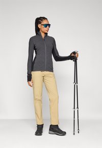 Black zip-up jacket with textured fabric, cream-colored lightweight pants, and black hiking boots. Holding black trekking poles, wearing blue sunglasses.