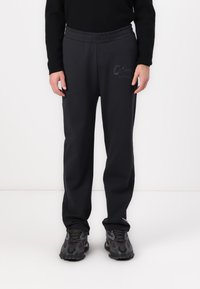Black sweatpants with an elastic waistband, featuring embroidered text on the left thigh, paired with textured black sneakers.