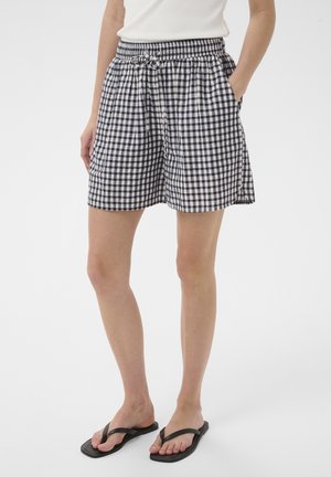 Nő fekete-fehér kockás, húzózsinóros rövidnadrágban, fehér ujjatlan felsőben és fekete papucsban, zsebeiben tartott kezekkel áll.