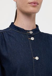 Temno modra jeans jakna s stoječim ovratnikom, srebrnimi gumbi in rumenim šivanjem. Opazna tekstura in krojena napihnjena manga dodajata podrobnosti.