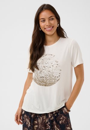 Donna con lunghi capelli castani sorride, indossa una maglietta bianca con disegno circolare astratto dorato e una gonna floreale scura, in piedi contro uno sfondo semplice.