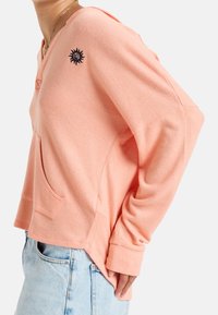 Sweat à capuche couleur pêche, coupe décontractée, poche avant, avec un motif de soleil brodé sur l'épaule droite. Tissu doux et texturé.