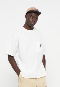 White cotton t-shirt with short sleeves and a chest pocket featuring a logo patch. Worn with light beige pants and a tan snapback cap.