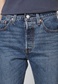 Jeans de denim azul con corte tapered, con una cinturilla de altura media, remaches de cobre y diseño clásico de cinco bolsillos. Textura desgastada visible.