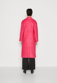 Cappotto di lana rosa con maniche lunghe, colletto a punta e taglio dritto. Morbida texture con una cucitura centrale che scende dalla schiena.