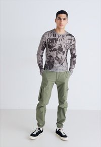 Camisa gris de manga larga con estampados gráficos, combinada con pantalones cargo holgados de color oliva que tienen bolsillos laterales y zapatillas negras con suelas blancas.