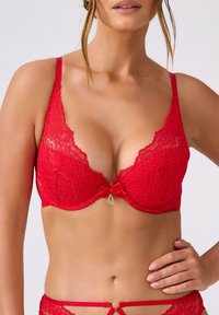 Czerwony koronkowy biustonosz z głębokim dekoltem, falistymi krawędziami, regulowanymi ramiączkami i małym złotym ozdobnym zawieszkiem na kokardce w centrum.