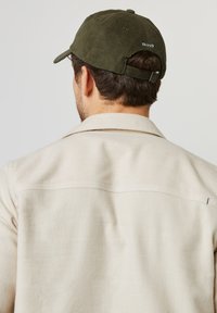 Mann mit einer cremefarbenen Jacke und einer olivgrünen Cordmütze, der von einem schlichten hellen Hintergrund weg schaut.