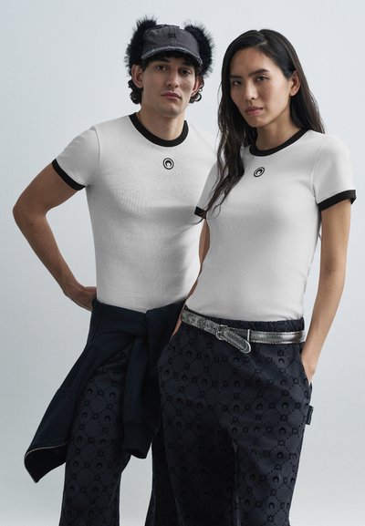 Deux mannequins portent des t-shirts blancs ajustés avec des cols ronds noirs et un logo. L'un porte un pantalon bleu marine à motifs, l'autre un sweat-shirt bleu marine.