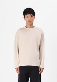 T-shirt beige à manches longues avec un col rond, fabriqué en coton doux, présentant une coupe décontractée et des détails de couture minimalistes.
