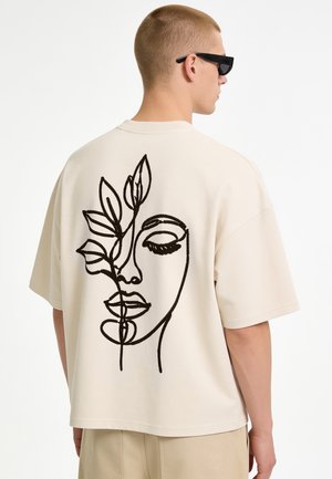 Young man wearing beige oversized t-shirt with black abstract face and leaf line art on the back, beige pants, and black sunglasses.