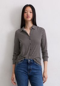 Camisa de botones de patrón marrón con cuello, mangas largas y un corte relajado, combinada con jeans azules, frente a un fondo neutral.