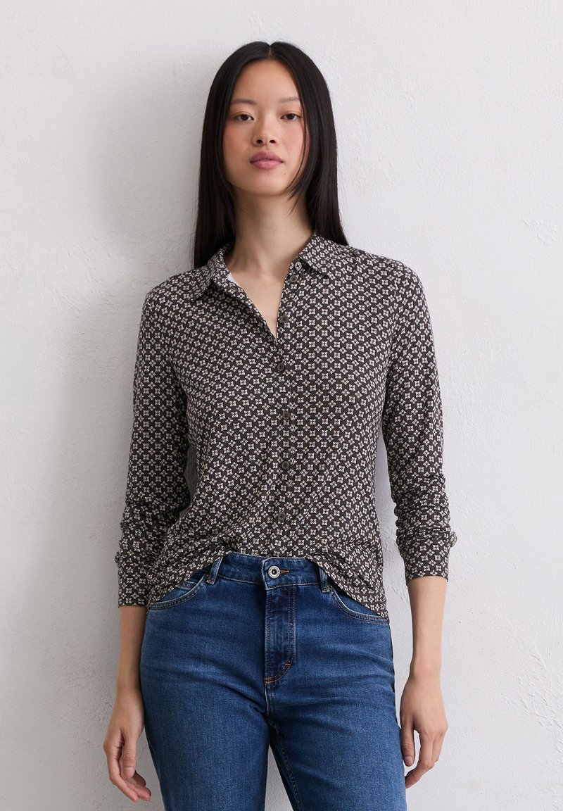 Camisa de botones de patrón marrón con cuello, mangas largas y un corte relajado, combinada con jeans azules, frente a un fondo neutral.
