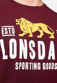 Bordo spalvos marškinėliai trumpomis rankovėmis su balta ir geltona grafiką, kurioje pavaizduota liūto emblema ir užrašas "ESTD LONSDALE SPORTING GOODS."