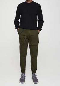 Zwarte geribbelde trui, olijfgroene cargobroek met zijzakken, trekkoord in de taille en grijze sneakers met lichte accenten.