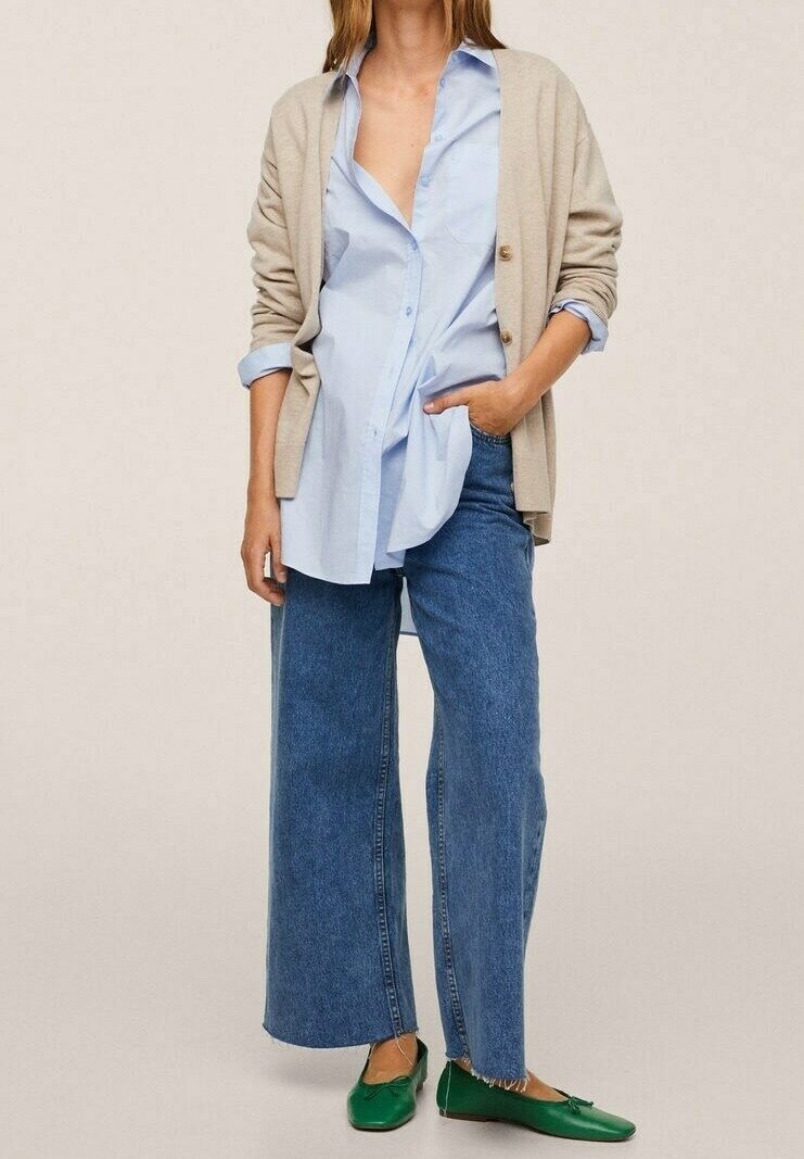 Beige Cardigan über einem hellblauen Hemd mit Knöpfen, kombiniert mit weiten blauen Jeans und grünen flachen Schuhen. Glatte Texturen und lässiger Schnitt.