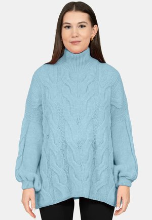 Junge Frau trägt einen hellblauen, grob gestrickten Rollkragenpullover mit langen, puffy Ärmeln und steht vor einem schlichten Hintergrund.