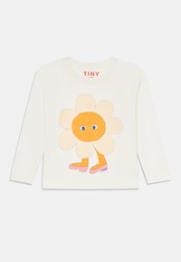 Valkoinen pitkähihainen paita, jossa on sarjakuvamainen kukka, jonka keskellä on oranssi, siniset silmät ja vaaleanpunaiset sandaalit, ja jossa on "TINY"-merkintä helmassa.
