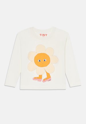 Hvit langermet skjorte med et tegneserieblomst med oransje sentrum, blå øyne og rosa sandaler, merket med "TINY" på kanten.