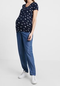 Navy blue maternity t-shirt with white leaf print, short sleeves, and a scoop neckline paired with loose-fitting blue denim jeans and white sneakers.