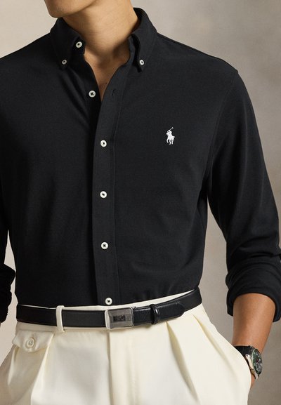 Μαύρο πουκάμισο μακρυμάνικο με �κουμπιά και λευκό λογότυπο, με αντίθετα κουμπιά, φορεμένο πάνω από μπεζ παντελόνια που στερεώνονται με μαύρη ζώνη.