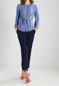 Žena v modré blůze s dlouhými rukávy a uzlem vpředu, tmavě modrých kalhotách a červených lodičkách, stojící s rukama v kapsách.
