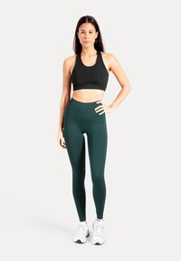 Reggiseno sportivo nero abbinato a leggings verde scuro; tessuto liscio, design aderente, vita alta e scarpe da ginnastica bianche.