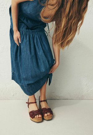 Pikade juustega naine, kellel on seljas varrukateta sinine kleit, kohendab serva, seistes punastes punutud platvormsandaalides tekstuurse seina taustal.