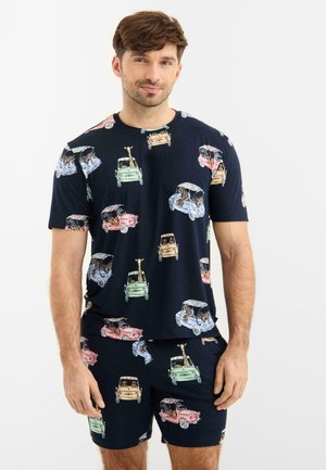 Mand iført matchende navyblå skjorte og shorts med farverige tegneserie-biler og dyrepassagerer på en hvid baggrund.