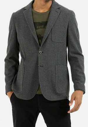 Homme portant un blazer gris texturé sur un t-shirt graphique vert olive et un pantalon noir, les mains partiellement dans les poches.