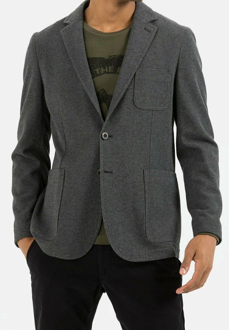 Homme portant un blazer gris texturé sur un t-shirt graphique vert olive et un pantalon noir, les mains partiellement dans les poches.