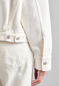 Bílá džínová bunda s texturovaným povrchem. Má stříbrné patentky na rukávu a kapsu na zadní straně, což zdůrazňuje minimalistický design.