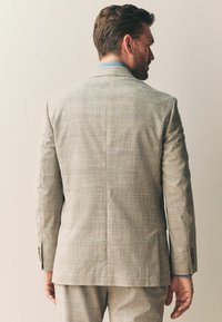 Chaqueta de traje gris claro con un patrón de cuadros. Presenta una abertura trasera única y detalles con botones marrones. Complementada con una camisa azul por debajo.