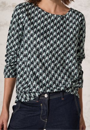 Mujer con blusa de manga larga con patrón de pata de gallo en negro, blanco y gris metida dentro de unos vaqueros azul oscuro.