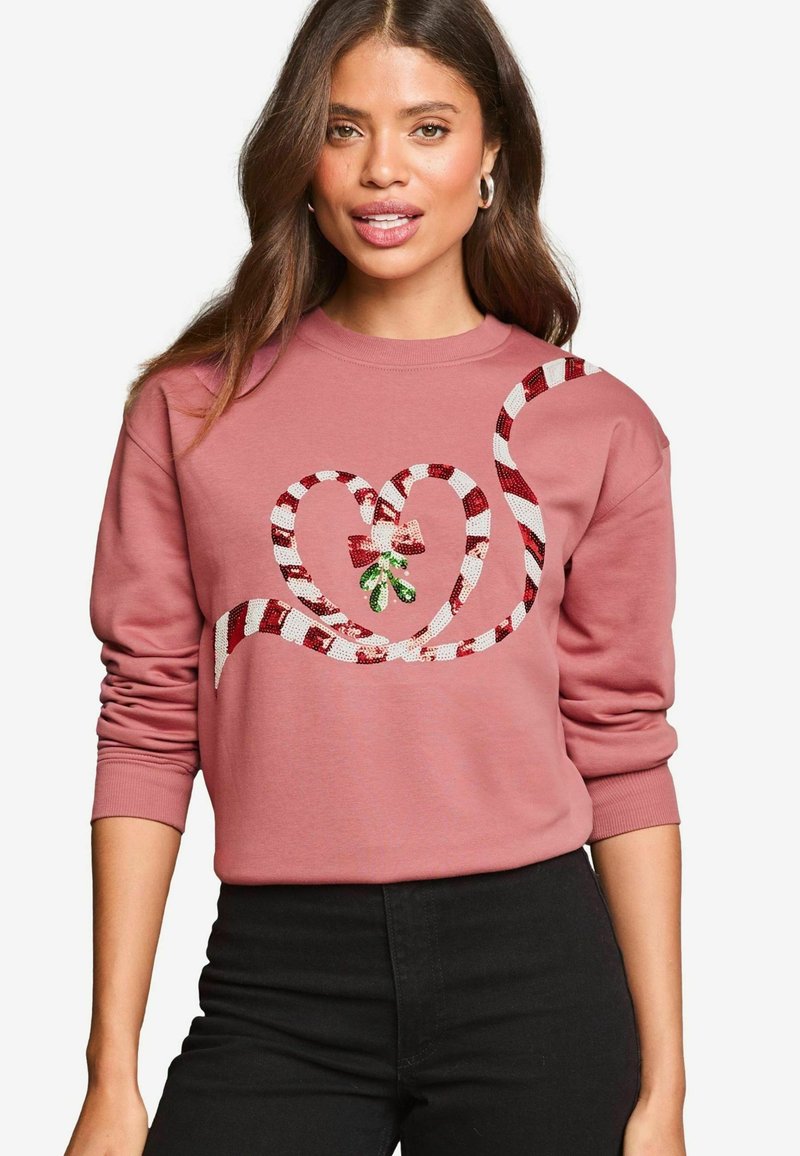 Sudadera rosa con un diseño en forma de corazón hecho de rayas de bastón de caramelo, con un acento de muérdago verde y un lazo rojo. Pantalones negros.