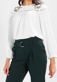 Blusa bianca con dettagli floreali ricamati sul colletto, maniche lunghe e tessuto testurizzato, abbinata a pantaloni verdi scuri a vita alta.