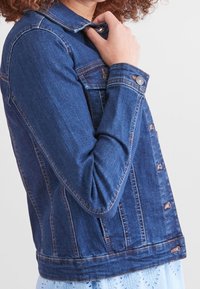 Chaqueta de mezclilla en azul oscuro con mangas largas, cierre de botones, bolsillos en el pecho y detalles de costura visibles. Llevada sobre una blusa de color claro.
