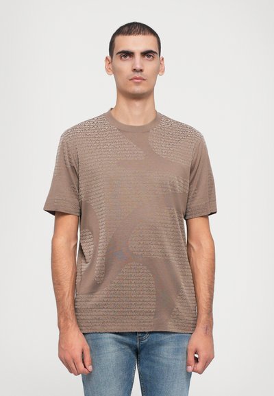T-shirt marron à manches courtes présentant un design texturé avec un logo répété, un col rond et une coupe décontractée. Porte avec un jean bleu.