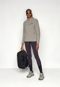 Pulover iz flisa svetlo sive barve s polovičnim zadrgo, kombiniran s vijoličnimi pajkicami in temnim nahrbtnikom. Model nosi sive pohodne čevlje in sončna očala.