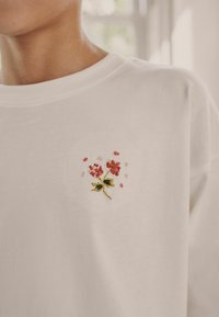 Weißer Baumwollpullover mit einem kleinen floralen Stickdesign in Rot und Pink mit grünen Stielen auf der linken Brustseite.