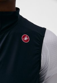 Svart ermeløs sykkelvest med høy krage, med en rund rød logo med en skorpion. Teksturert hvit lag synlig under.