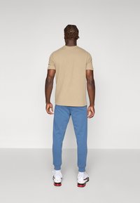 Beige t-shirt med kort ärm, texturerat tyg; blå träningsoverallsbyxor med åtsittande anklar; vita sportskor med svarta accenter.