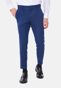 Pantaloni su misura blu con fronte piatto, tasche frontali e risvolti, abbinati a mocassini neri e calzini neri. Texture del tessuto liscia.