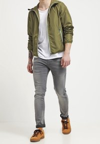 Olivgrön lättjacka med huva, vit T-shirt, grå slim-fit jeans och bruna sneakers med bruna skosnören.