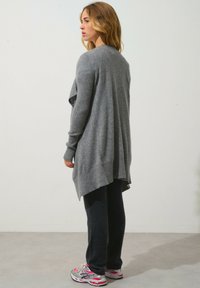 Cardigan lavorato a maglia grigio chiaro con una vestibilità ampia, orlo asimmetrico e polsini a coste, abbinato a pantaloni grigio scuro e sneaker sportive.