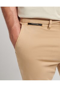 Pantalones beige con una textura de tela suave, que presentan una etiqueta de marca negra en la cintura y bolsillos laterales. Ajuste entallado.