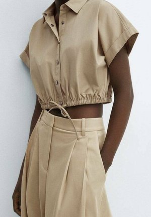 Personne à la peau foncée portant une chemise beige courte à boutons avec taille coulissante et un pantalon plissé taille haute, sur fond blanc.
